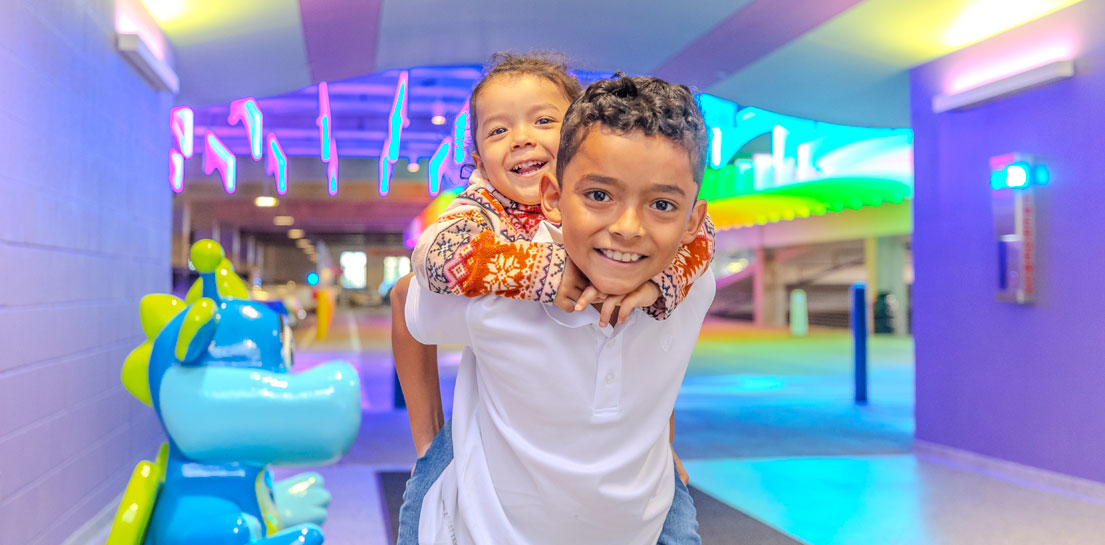 Smiling boy gives piggyback ride to a child in a brightly lit, colorful clinic hallway with neon art and a blue statue