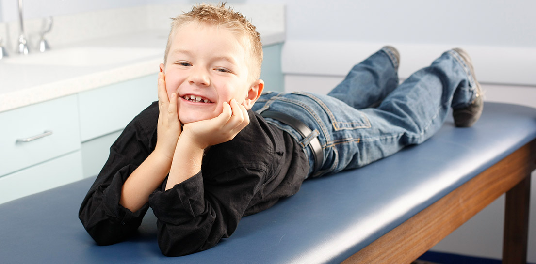 Young boy smiling on a blue exam table, resting chin on hands